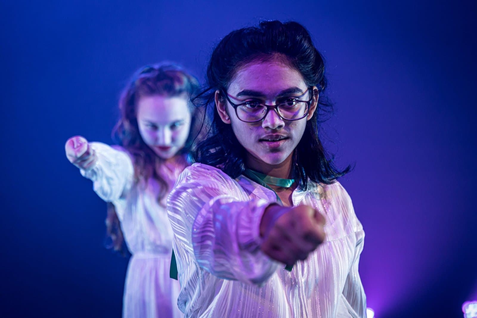 Two performers reaching forward under blue and violet stage light.